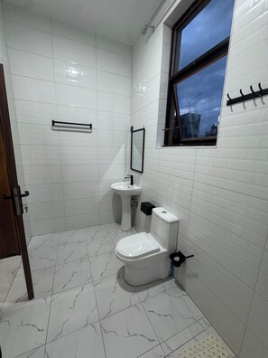 Ein Badezimmer mit weißen Fliesen, einer Toilette, einem Waschbecken und einem Fenster mit Blick auf den Himmel.
