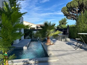 Una piscina con borde de baldosas, rodeada de vegetación y una pérgola.