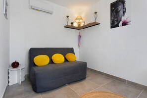 Ein Wohnzimmer mit grauem Sofa, drei gelben Kissen, einem kleinen weißen Beistelltisch mit einem Apfel, einer Wand-Klimaanlage, einem Wandregal mit einer Lampe und einem gerahmten Bild.