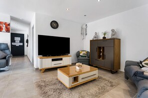 Ein modernes Wohnzimmer mit Flachbildfernseher, Couchtisch aus Holz, grauem Sofa und einer Kommode mit dekorativen Gegenständen.
