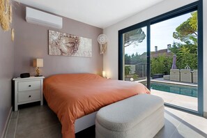 A bedroom with a large bed, a bedside table, a lamp, a wall-mounted air conditioner, and a view of a swimming pool through a large window. - Villa Harmonie with Private Pool, Wi-Fi, and Air Conditioning (Bagnols-sur-Cèze)