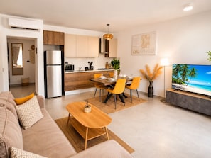 Ein modernes Wohnzimmer mit Sofa, Couchtisch, Essbereich, Küche und Fernseher, der eine Strandansicht zeigt.