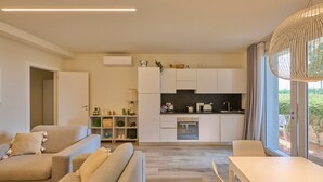 Una sala de estar moderna con un sofá y una mesa de comedor, una cocina con armarios y electrodomésticos blancos, y un ventanal con vistas.