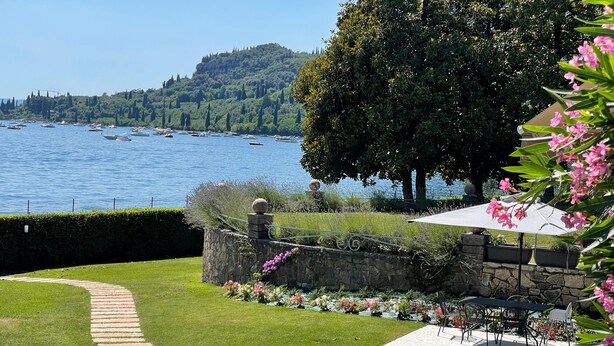 Jardim ajardinado com muro de pedra, caminho pavimentado e vista para barcos na água.