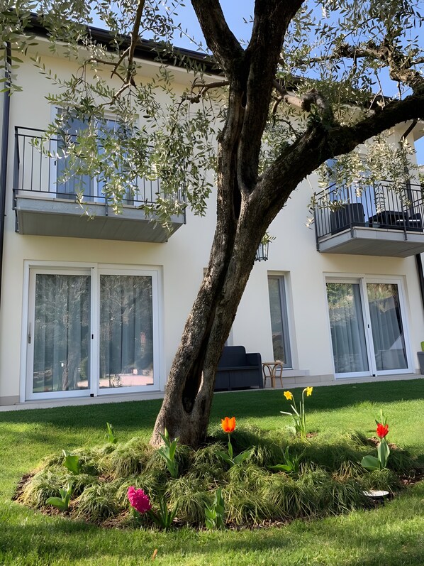 A residential building with a large tree in the foreground, a balcony with a railing, and a grassy lawn with flowers. - Apartment 'Flora' with Lake View, Wi-Fi and Air Conditioning (Garda)