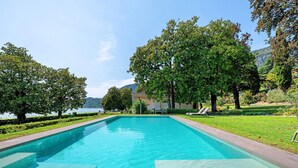 A rectangular swimming pool with clear blue water, surrounded by well-maintained grass and trees, with a view of a lake and hills in the background. - Apartment 'Matilde' with Lake View, Wi-Fi and Air Conditioning (Garda)