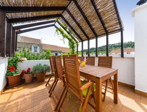 Eine überdachte Terrasse im Freien mit Tisch und Stühlen, Blumentöpfen und Blick auf ein Gebäude mit Ziegeldach.