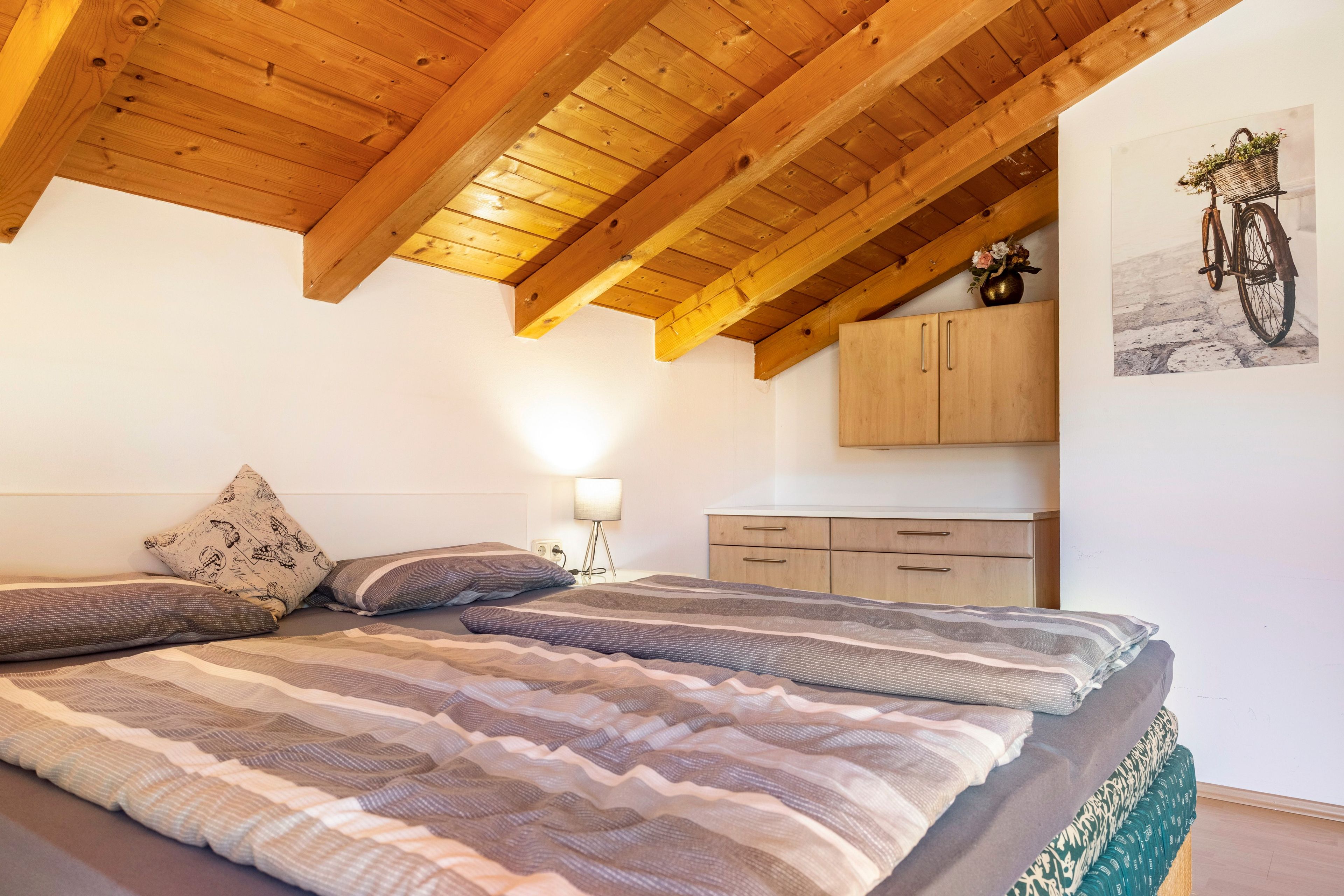 A bedroom with a wooden ceiling, a bed with striped bedding, a nightstand with a lamp, and a wall-mounted cabinet.