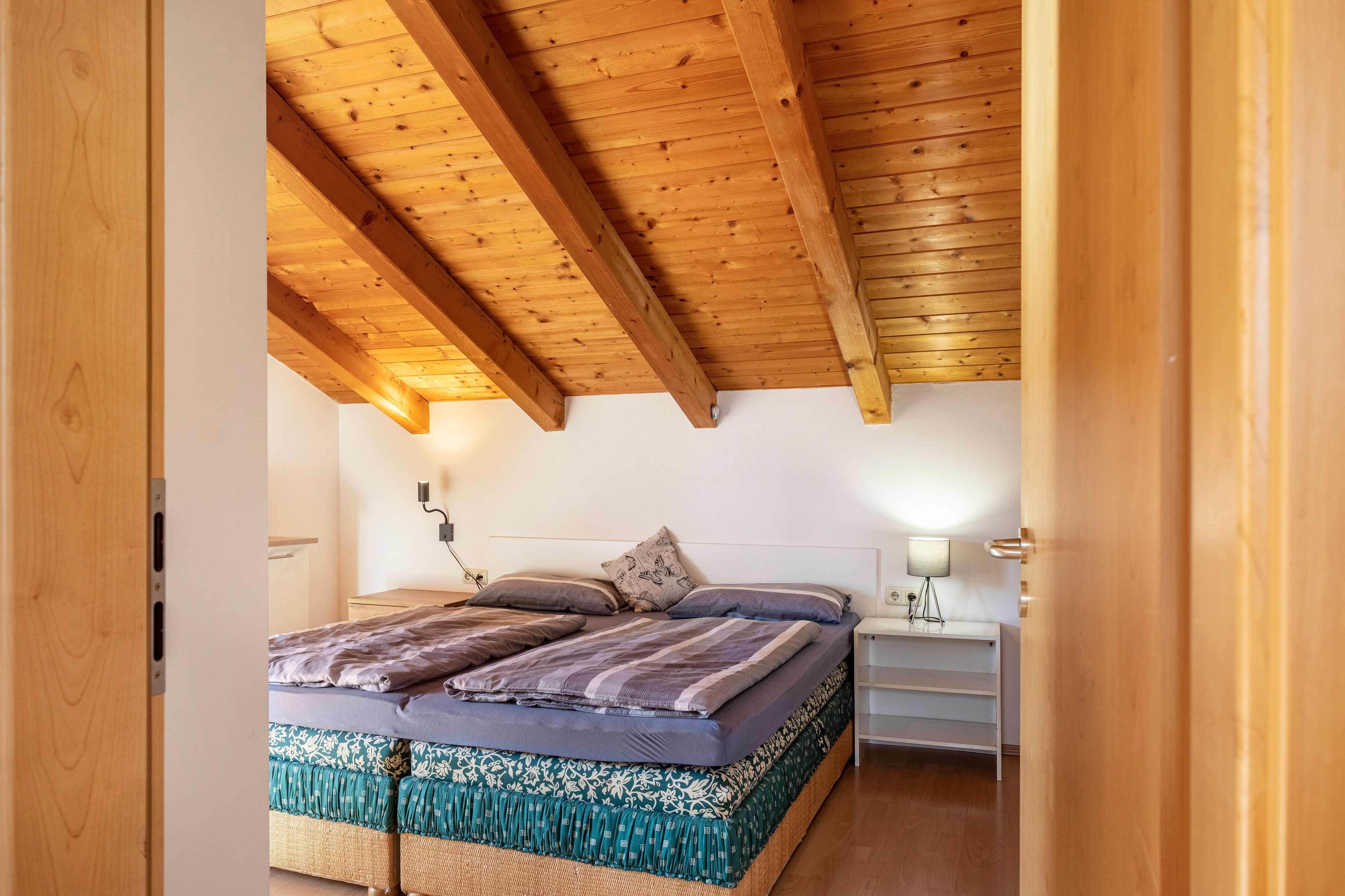 Ein Schlafzimmer mit Holzboden, einem Bett mit gemusterten Betttextilien, einem Nachttisch mit Lampe und einem Beistelltisch.