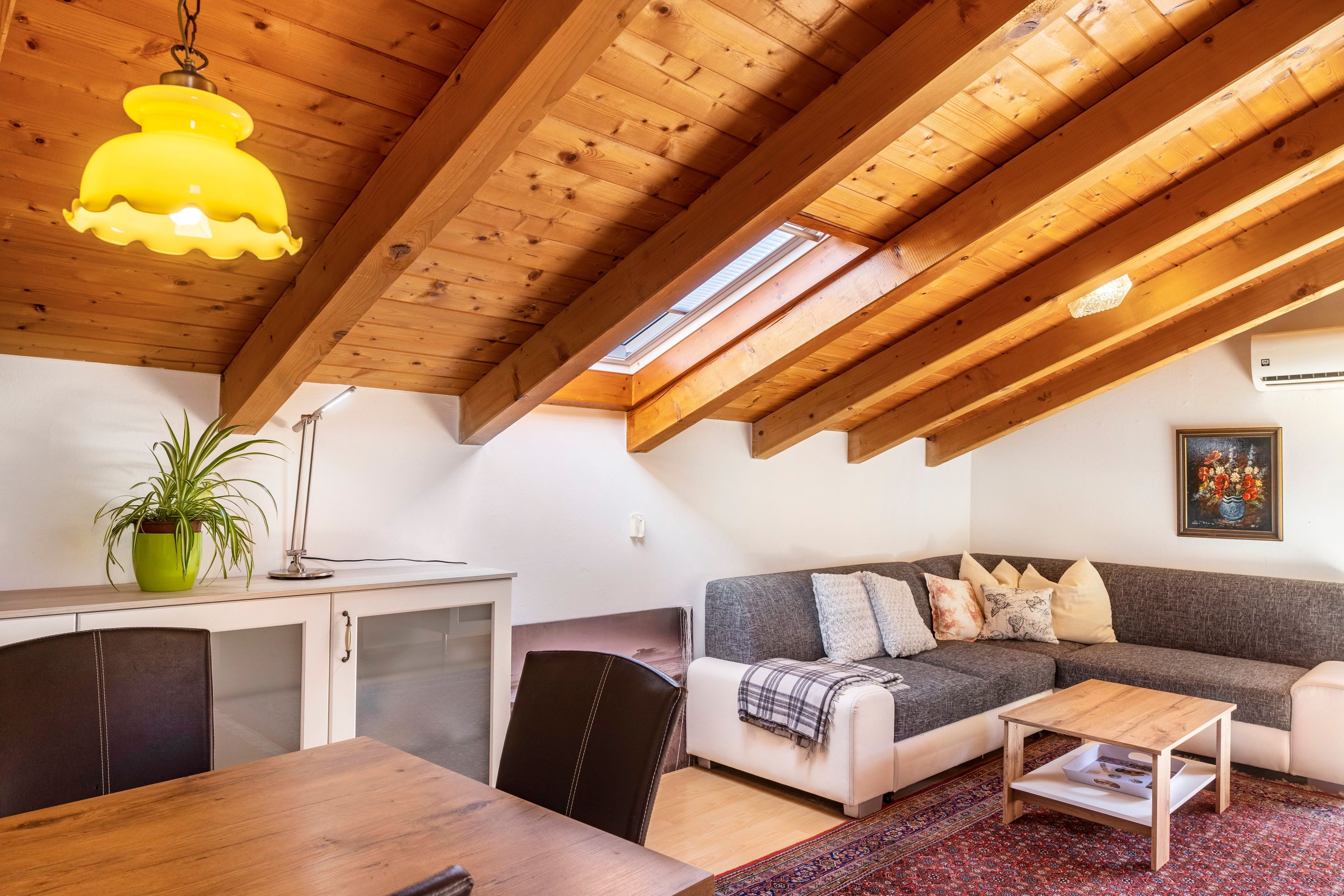 Ein gemütliches Loft-Zimmer mit Holzbalkendecke, einem Esstisch, einer Couch und einer Topfpflanze.
