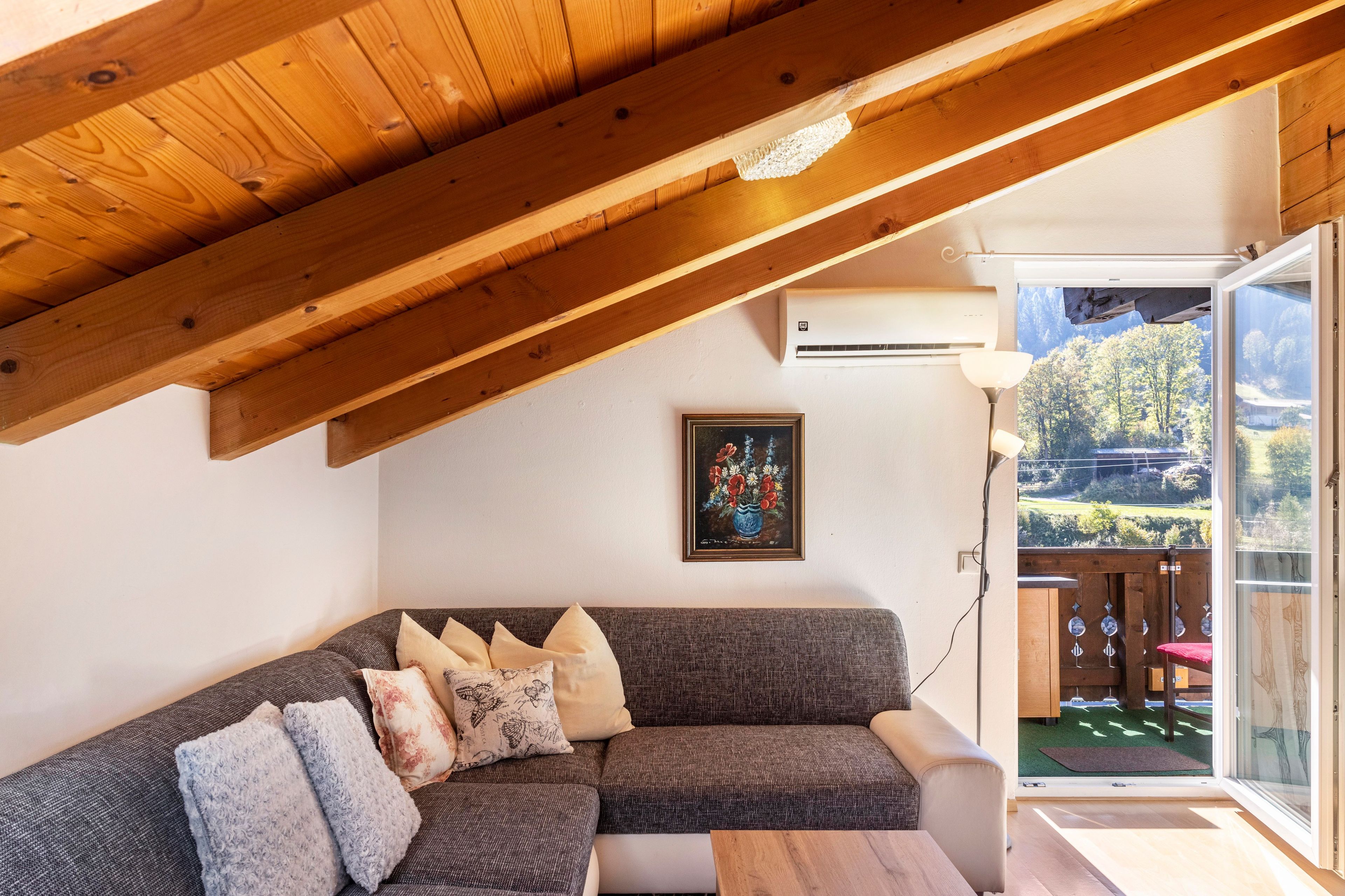 Ein gemütliches Wohnzimmer mit Holzboden, einer Couch mit gemusterten Kissen, einem gerahmten Kunstwerk und Blick auf einen Garten durch eine Glastür.