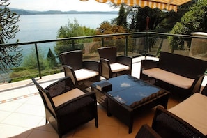 A balcony with wicker furniture and a glass table, overlooking a lake.