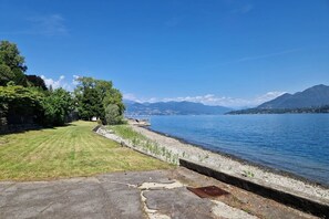 A lakeside view with a grassy area, a concrete path, and mountains in the background.