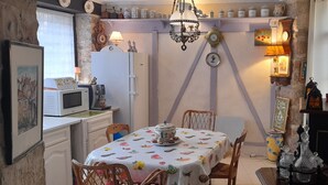 Ein gemütlicher Essbereich mit einem Tisch für zwei Personen, umgeben von Korbstühlen. Eine Chandelier hängt darüber, und verschiedene Küchengeräte sowie Dekorationsgegenstände sind sichtbar.