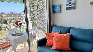 Un balcón con una mesa, sillas y un sofá azul con cojines naranjas. Hay plantas en una celosía y obras de arte en las paredes.