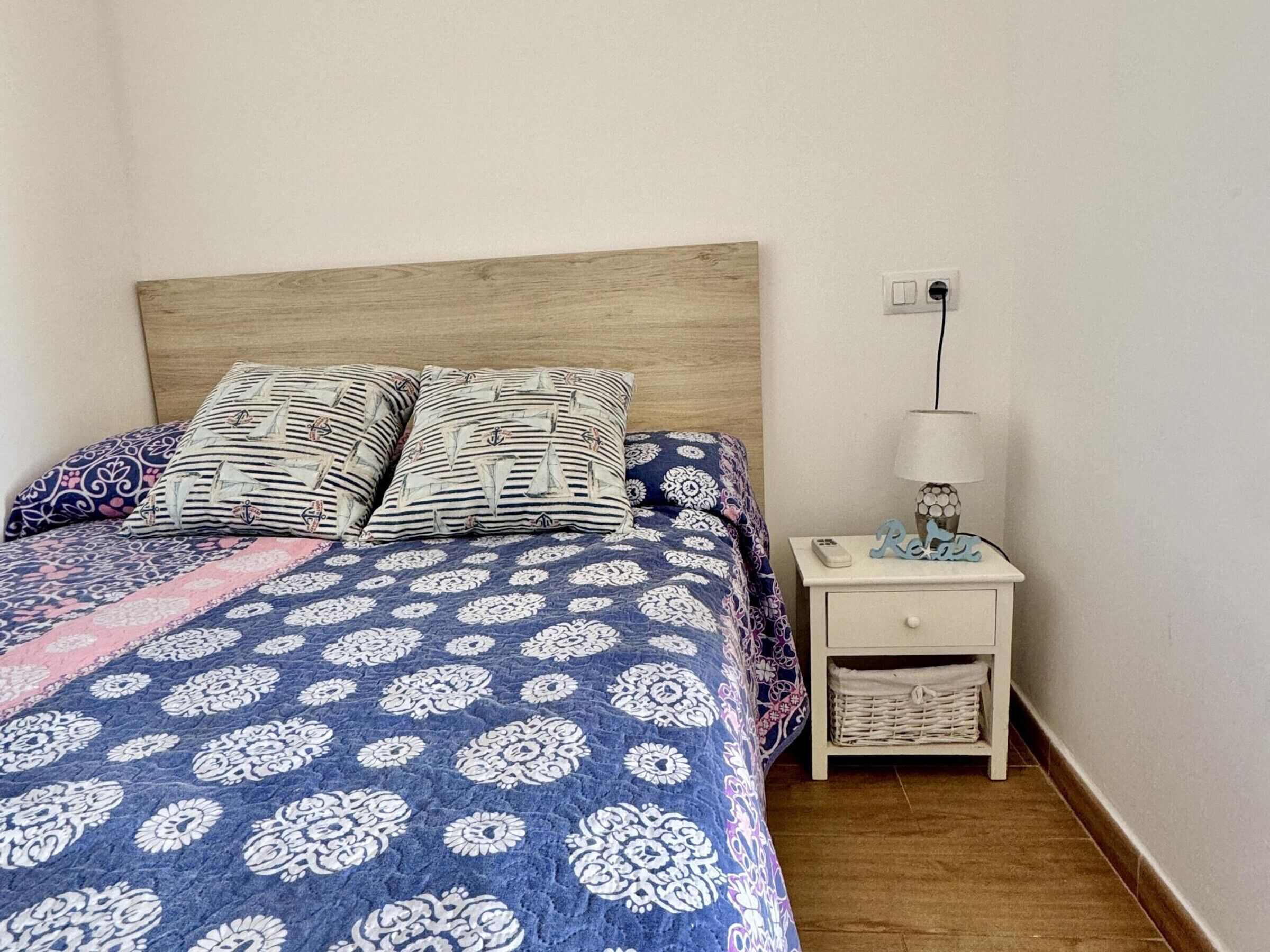 Een slaapkamer met een bed, twee kussens, een nachtkastje met een lamp en een klein opbergrek.