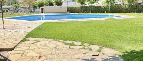 Une piscine avec une terrasse en pierre et de l’herbe au premier plan, entourée de bâtiments.