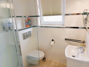 Ein Badezimmer mit Toilette, einer Glasdusche, einem Waschbecken inklusive Spiegel und einem Fenster mit Jalousien.