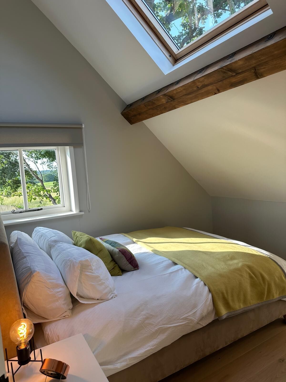 A bedroom with a skylight, a bed with pillows, a bedside lamp, and a window with a view of greenery.