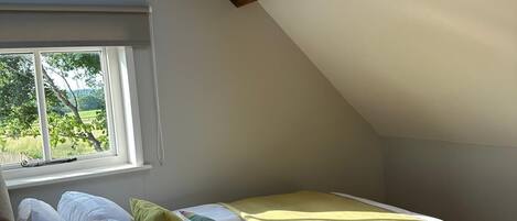 A bedroom with a skylight, a bed with pillows, a bedside lamp, and a window with a view of greenery.