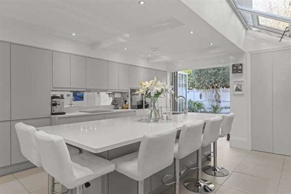 A modern kitchen with white cabinetry, a central island with bar stools, and a view of the outdoors. - Stylish Windsor Home | Hot Tub | Town Centre (Windsor)