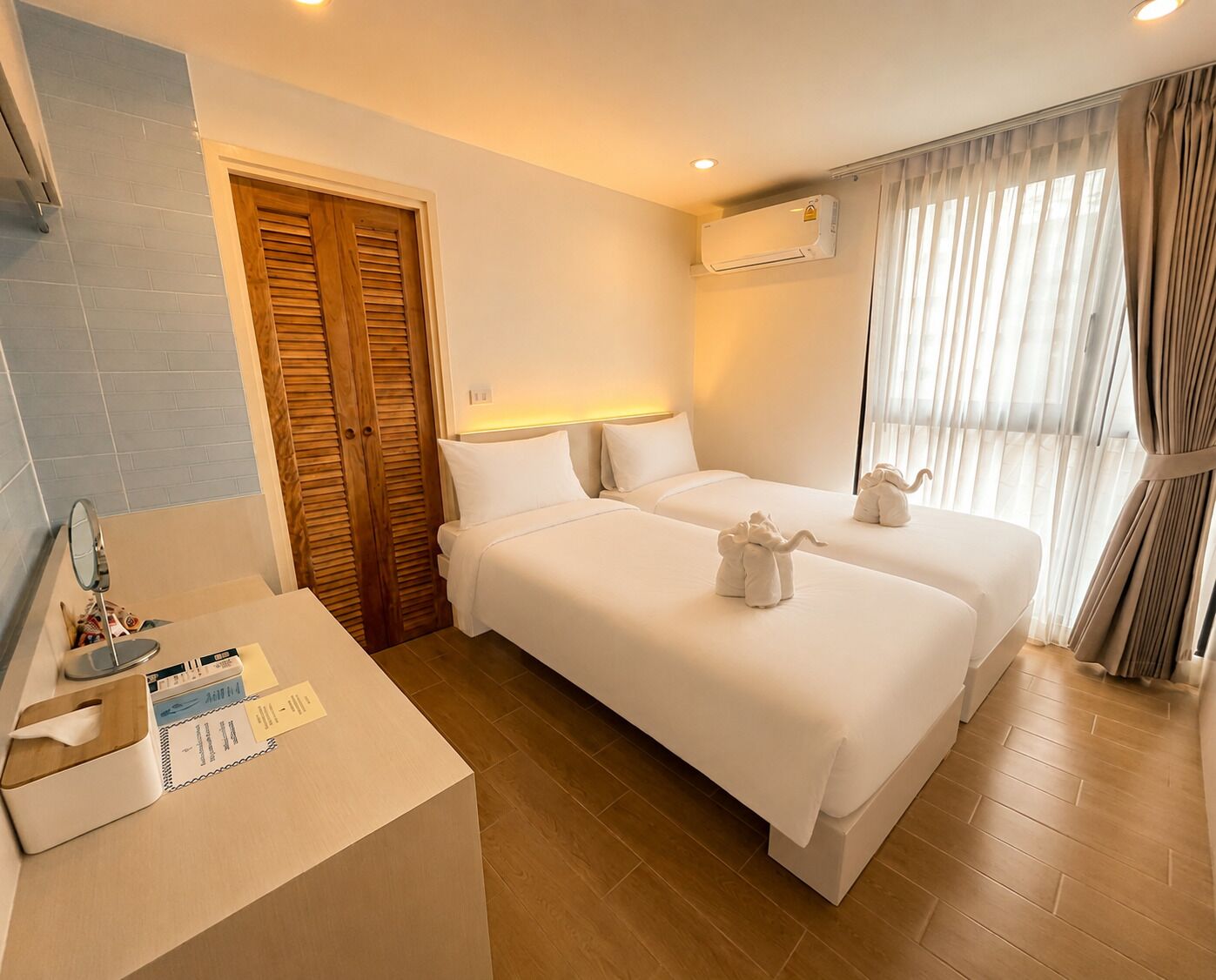A hotel room with two beds, a wooden door, an air conditioning unit, a bedside table with a mirror and toiletries, and a window with curtains.