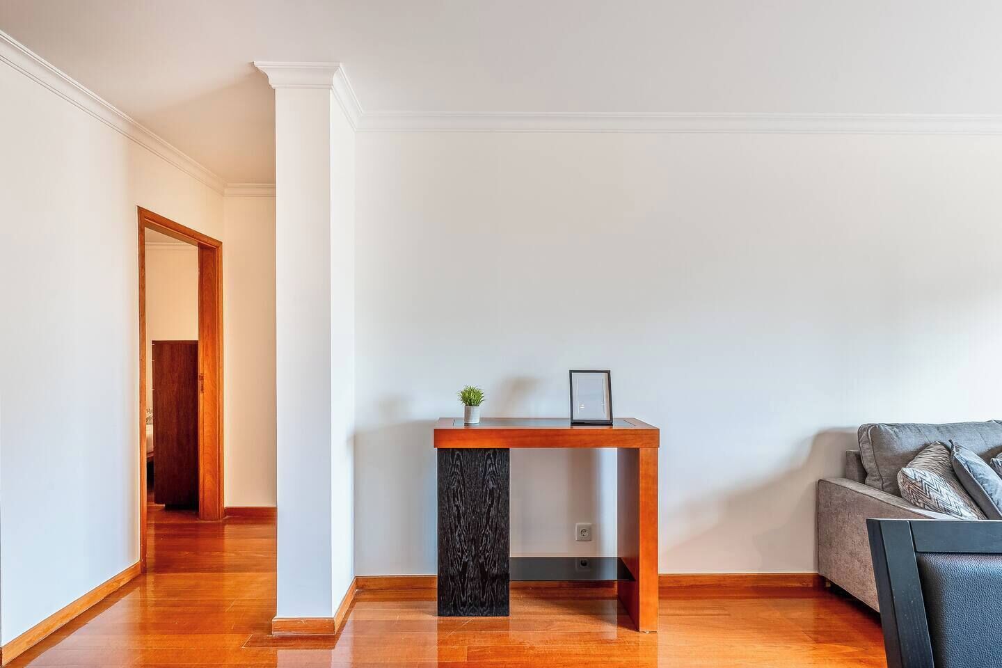 A room with a wooden floor, a dark wood table with a small plant and picture frame, a grey sofa, and a white wall with a door leading to another room.