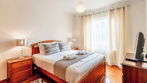 Ein Schlafzimmer mit einem hölzernen Bett, Nachttischlampen, einem Schreibtisch mit Computer und einem Fenster mit Vorhängen.