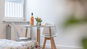 A room with a bed, a small table with a bottle and flowers, a chair, and a radiator.