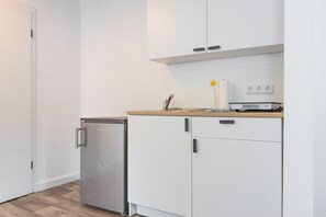 Eine kompakte Küche mit weißen Schränken, einem kleinen Kühlschrank und einer Arbeitsplatte mit Spüle und Lampe.