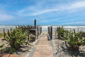 A pathway leading to a beach with a fence, plants, and a trash bin.