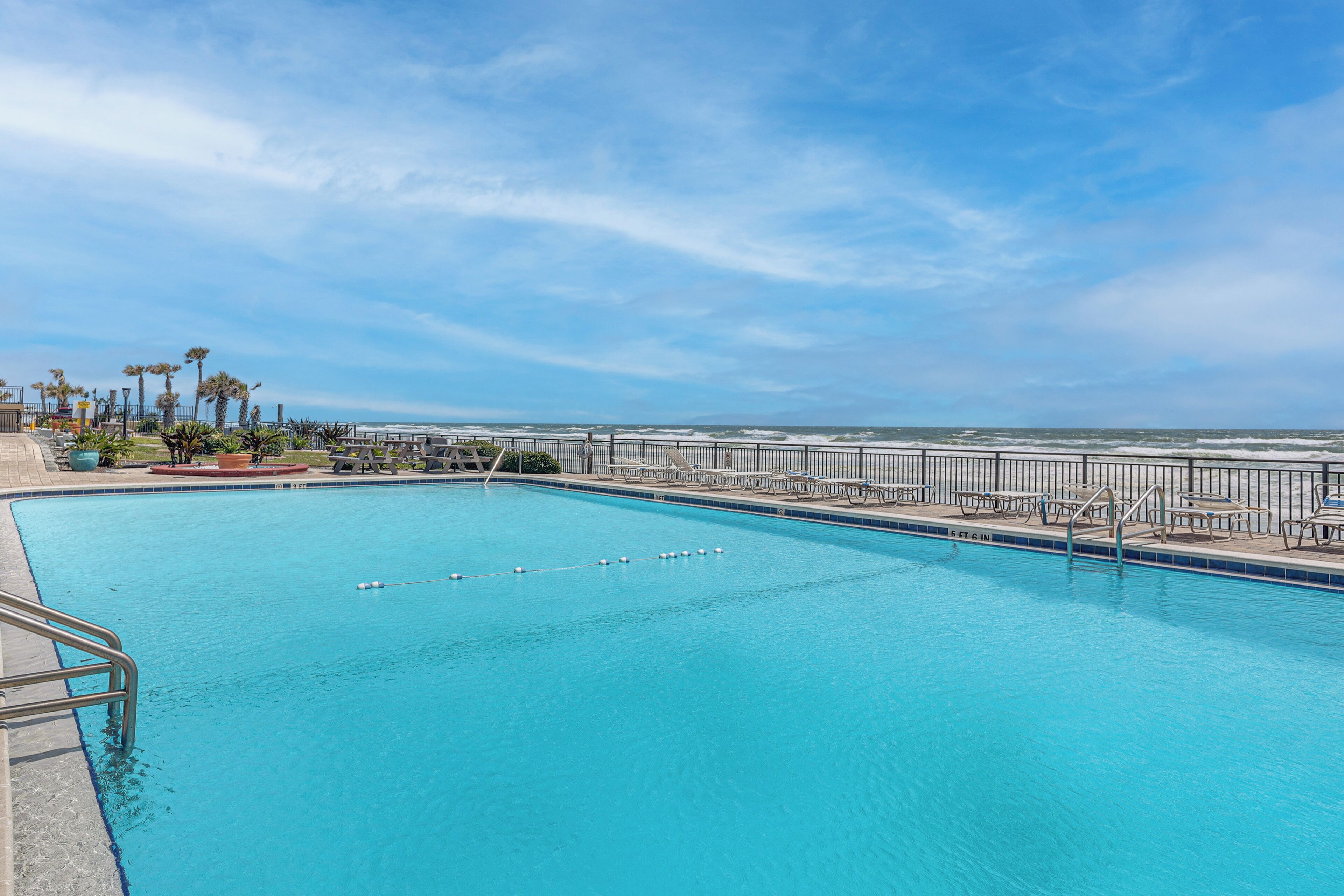 A swimming pool with a view of the ocean, surrounded by a fence and lounge chairs.
