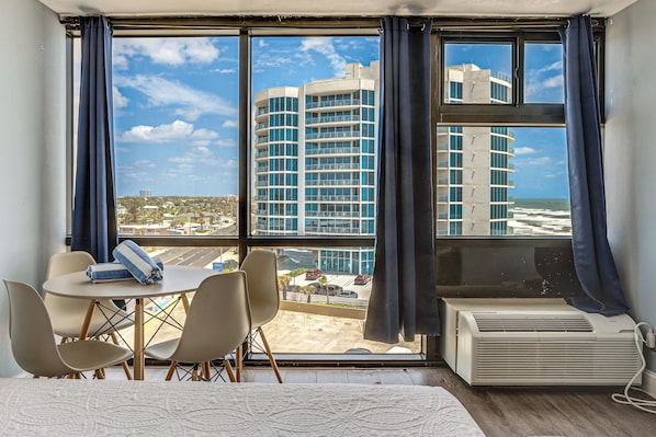 A hotel room with a large window, a table with chairs, a bed, and an air conditioning unit.