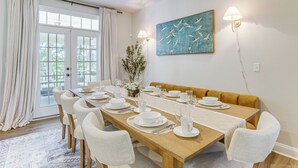 A dining room with a long table set for six, a sofa, and a painting on the wall.