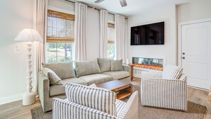 A living room with a sofa, armchairs, a coffee table, a fireplace, a ceiling fan, and a TV mounted on the wall.