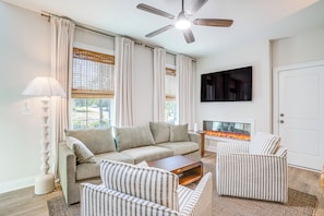 A living room with a sofa, armchairs, a coffee table, a fireplace, a ceiling fan, and a TV mounted on the wall.