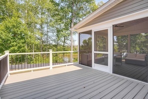 A covered outdoor deck with a railing, a glass door leading to an outdoor seating area, and a view of trees and a body of water.