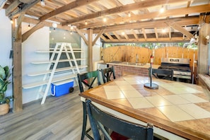 A covered outdoor dining area with a wooden table, chairs, a grill, and a ladder.