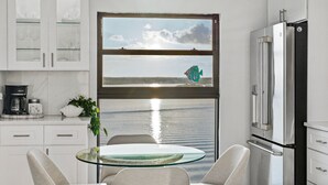 Een moderne keuken met een glazen tafel, witte kasten en uitzicht op zee door een raam.