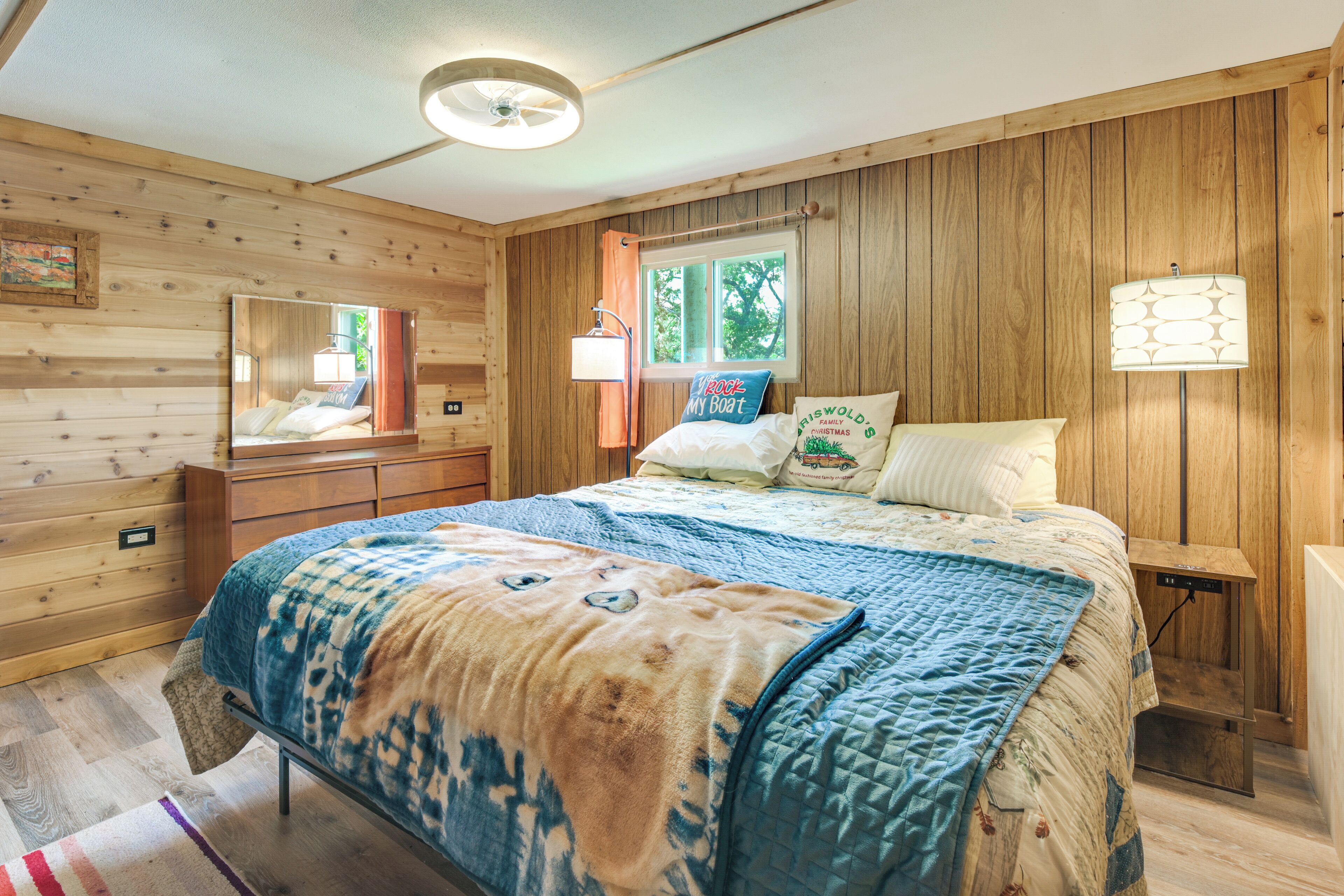 A wooden cabin bedroom with a bed, dresser, mirror, and window.