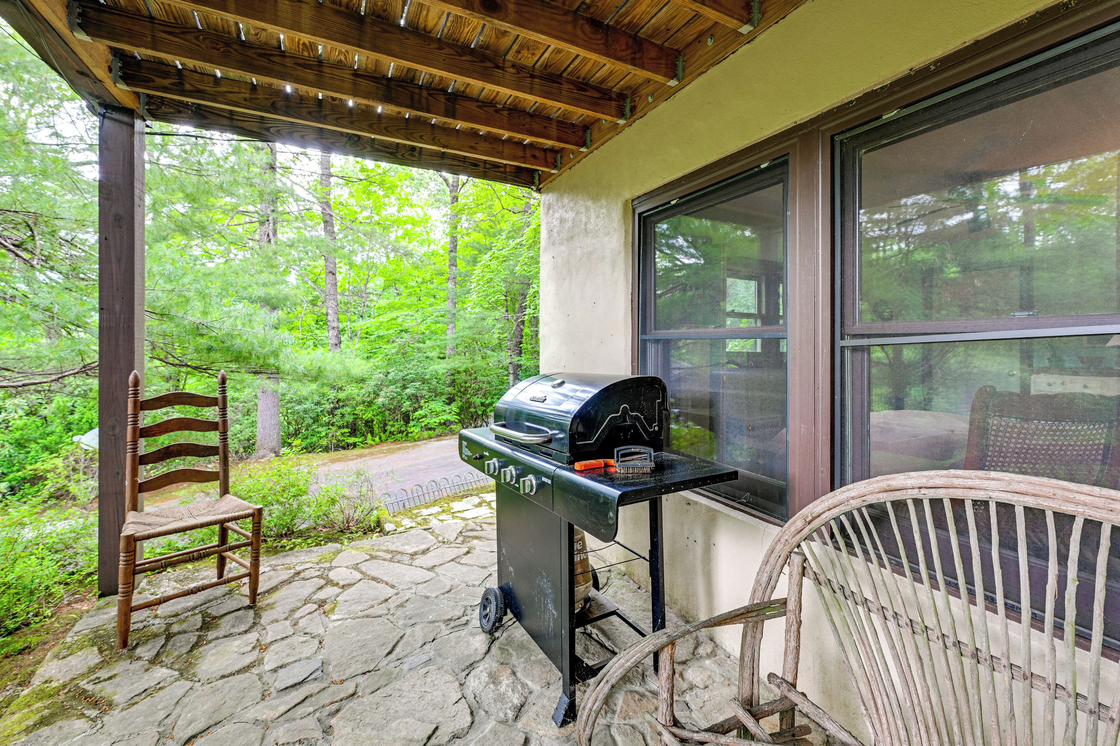 A covered patio with a stone floor, a grill, a wooden chair, and a wicker chair.