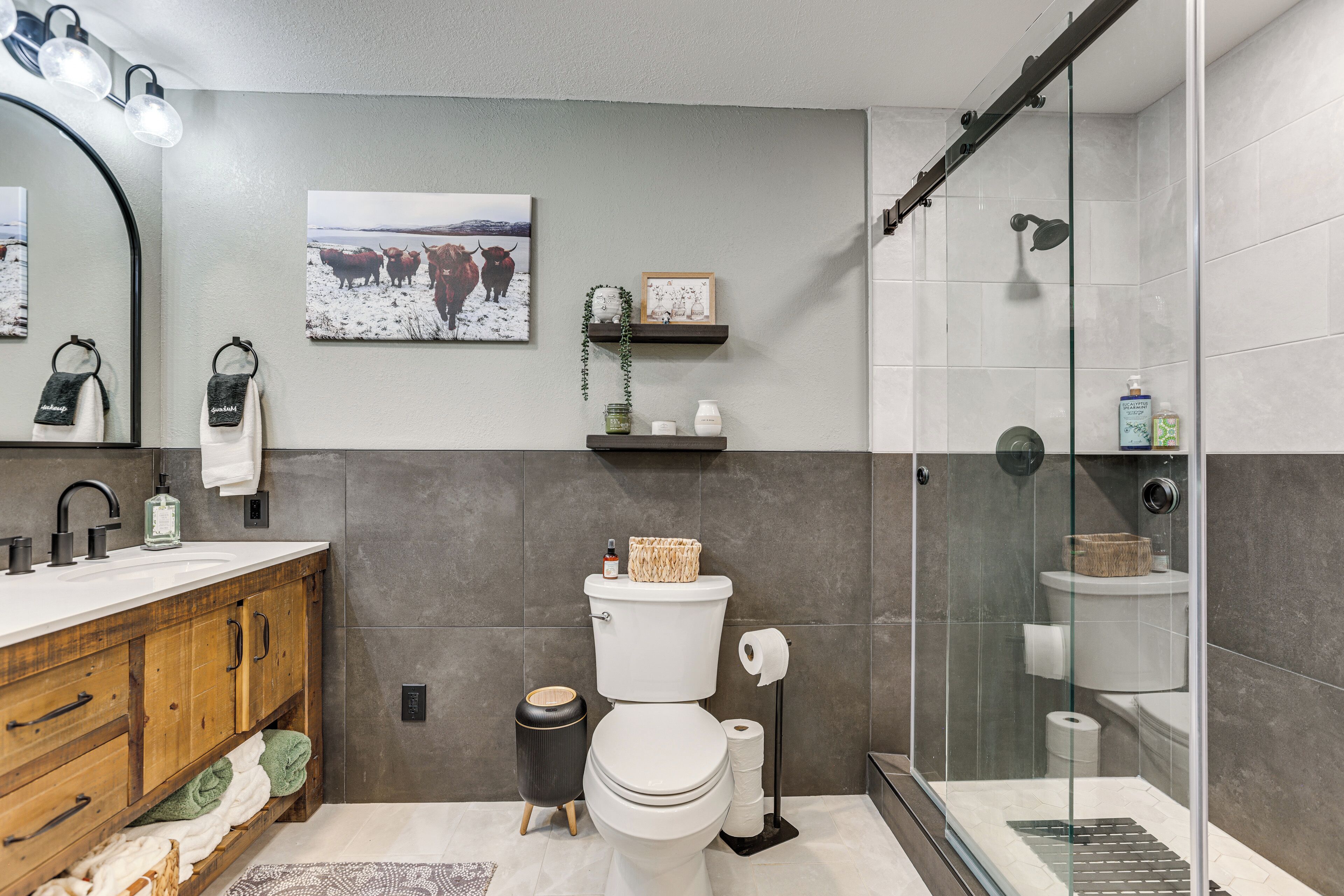 A bathroom with a wooden vanity, a toilet, a glass shower enclosure, and a wall-mounted shelf with toiletries.