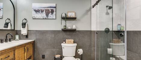 A bathroom with a wooden vanity, a toilet, a glass shower enclosure, and a wall-mounted shelf with toiletries.