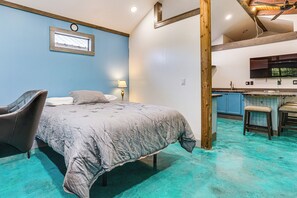 A bedroom with a bed, a chair, a desk, and a kitchenette.
