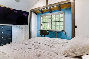 A bedroom with a bed, a desk, a chair, a TV mounted on the wall, and a window with a view of trees.