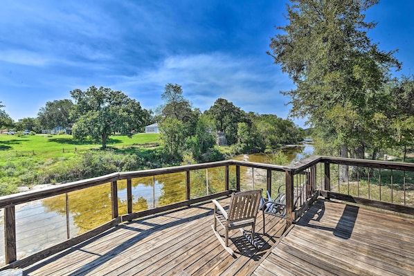 A wooden deck with a bench overlooking a river and a grassy area with trees.