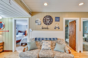 A cozy living area with a large clock, a couch with patterned cushions, and a wall with framed photos and a 'beach' sign.
