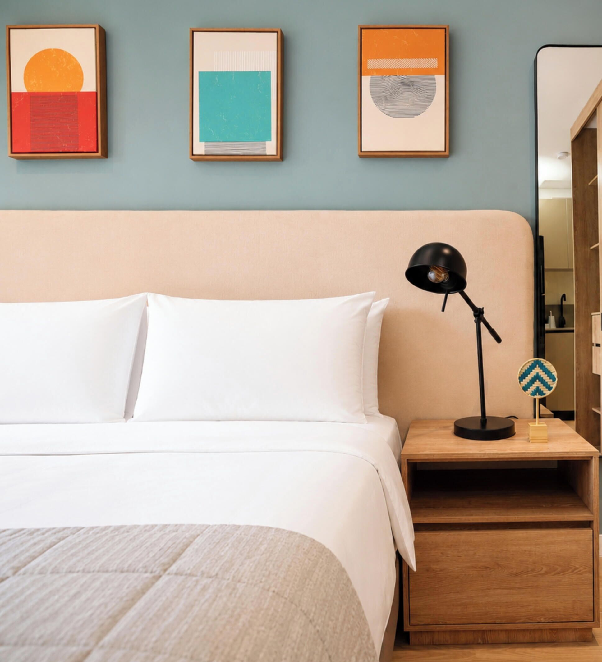 Una cama bien hecha con ropa de cama blanca, una mesita de noche de madera con una lámpara, y tres cuadros enmarcados en la pared.