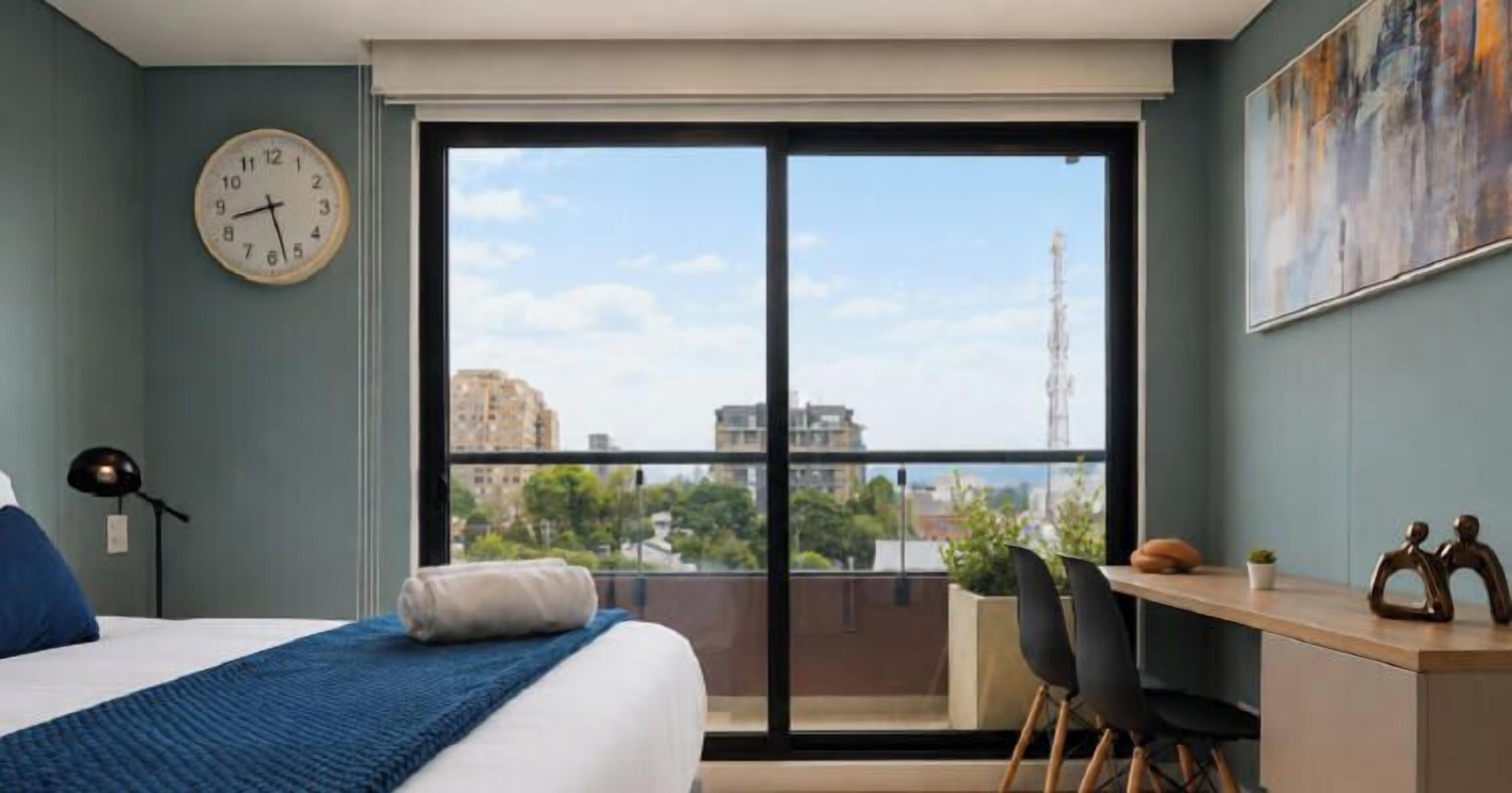 Habitación de hotel con cama, escritorio, silla, reloj y un amplio ventanal con vista a la ciudad.