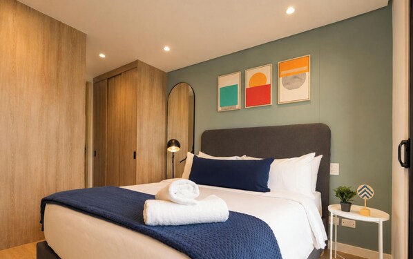 A neatly made bed with a blue blanket and white pillows, a bedside table with a plant, and a wall with three framed artworks. - WELL 100 by Jalo (Bogotá)